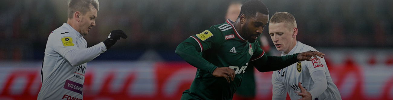 Футбольный матч: игрок в зелёной форме с мячом против двух соперников в белой форме. На заднем плане видны трибуны с небольшой размытостью