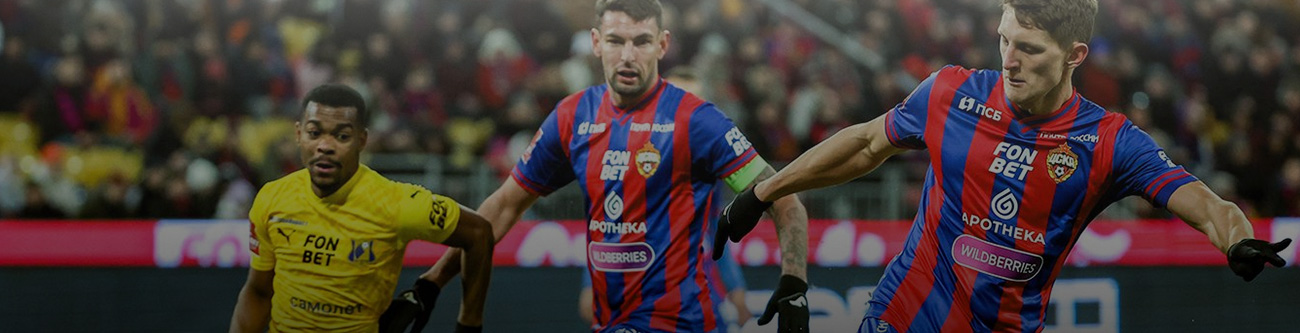 На изображении футболисты в разгаре игры: игрок в жёлтой форме против двух игроков в красно-синей форме на стадионе. На заднем плане видны трибуны с болельщиками