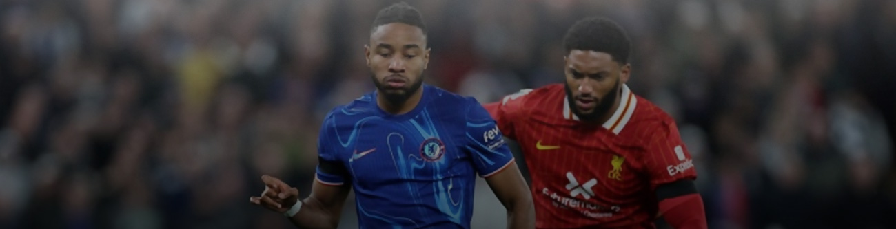 На изображении запечатлен момент футбольного матча: два игрока соперничают за мяч. Один в синей форме, другой в красной. На фоне видна размытой трибуна с болельщиками