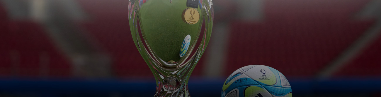 На изображении крупный план трофея и футбольного мяча на фоне размытого красного стадиона