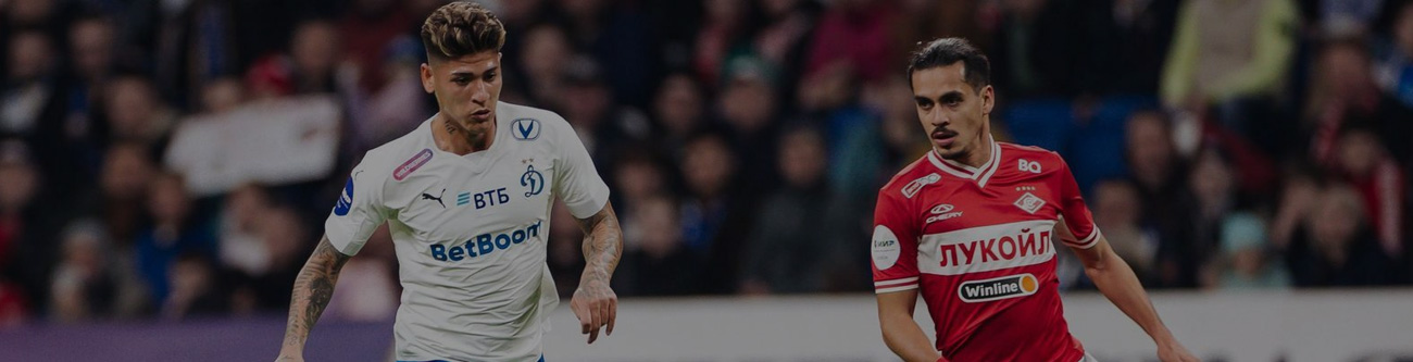 На изображении два футболиста на поле во время матча. Игрок в белой форме с логотипом «ВТБ» противостоит игроку в красной форме с надписью «Лукойл». На заднем плане видны болельщики