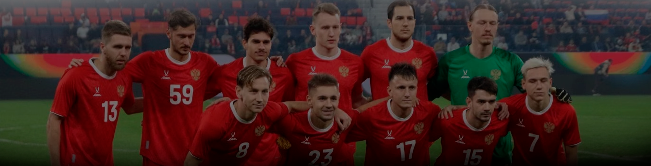 Футбольная команда в красной форме позирует для группового фото на поле перед матчем. Игроки стоят в два ряда на фоне трибун стадиона