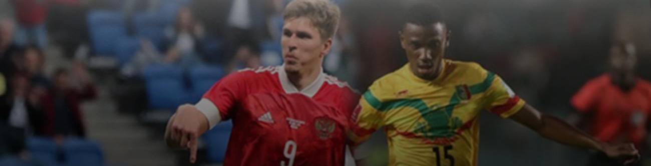 На изображении два футболиста в движении на поле: один в красной форме с номером 9, другой в желтой форме с зелеными и красными элементами. На заднем плане видны зрители на трибунах