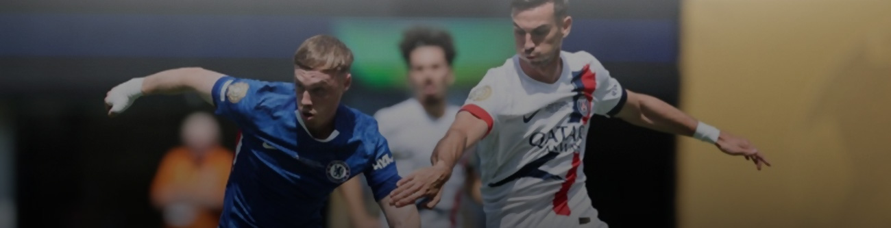 На изображении запечатлен футбольный матч: игрок в синей форме противостоит игроку в белой форме с красно-синим элементом. Они активно борются за мяч. На заднем плане виден размытый фон и другие участники