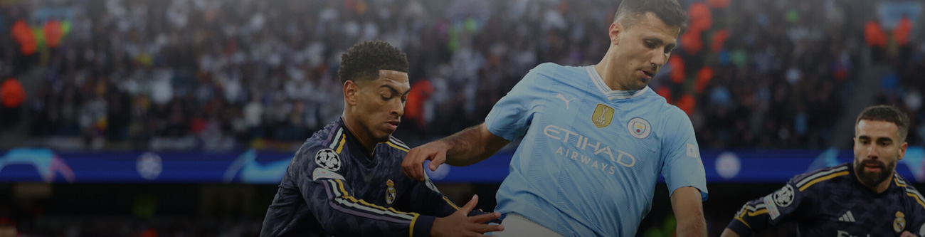 Футболист в голубой форме пытается обыграть соперника в тёмной форме на фоне зрителей на стадионе. На игроках видны клубные эмблемы, а игра, вероятно, проходит в рамках международного турнира