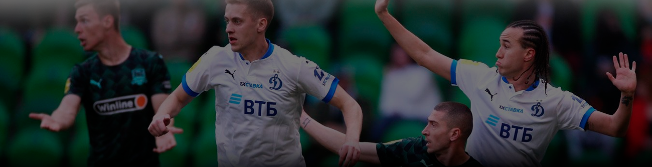 На изображении запечатлен момент футбольного матча. Игроки в белой и темно-зеленой форме борются за мяч. Видно активное противоборство на поле и напряженную игровую ситуацию