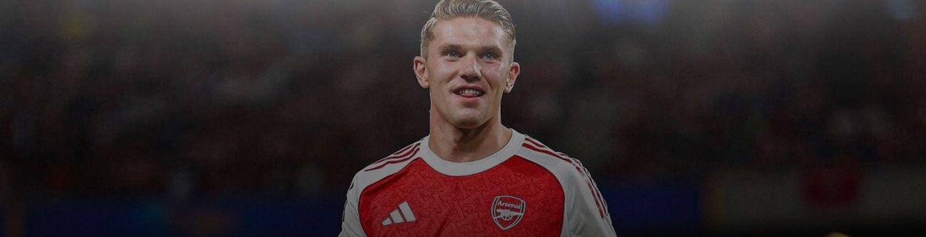 Молодой мужчина в красно-белой футболке футбольной команды с логотипом «Arsenal» находится на фоне размытой спортивной арены, освещенной прожекторами