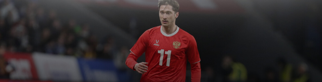 Футболист в красной форме с номером 11 на груди на игровом поле. На заднем плане видны размытые трибуны с болельщиками и участками флага