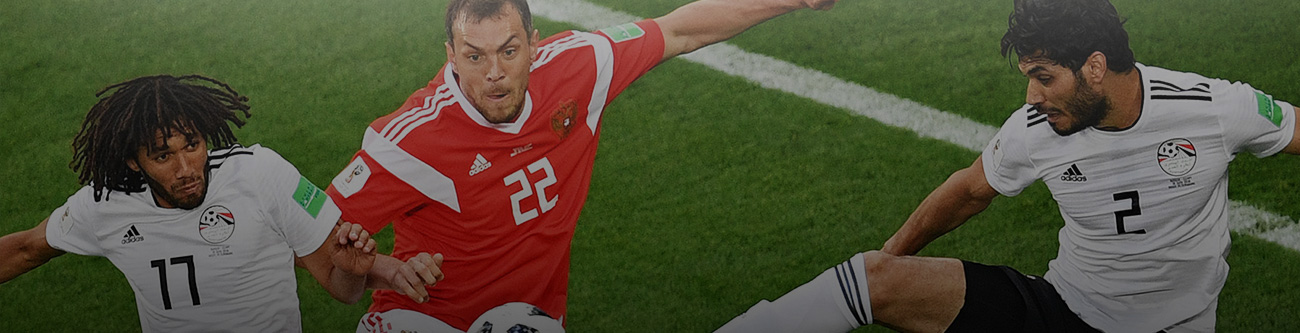 На изображении три футболиста борются за мяч на поле. Один в красной форме, двое других в белой. Динамичная игра, действие происходит на зеленом газоне